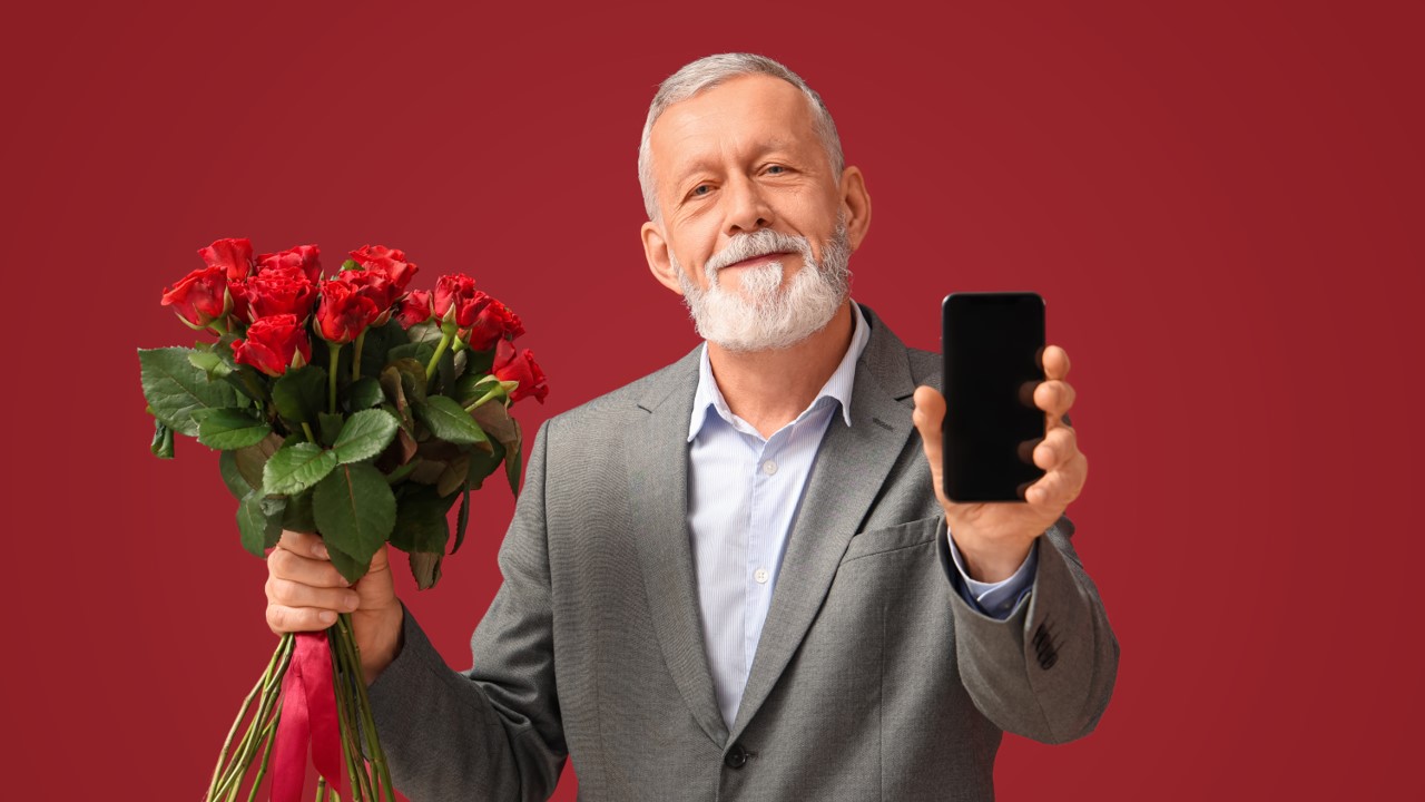 Homem mais velho sorrindo e segurando flores enquanto aponta celular